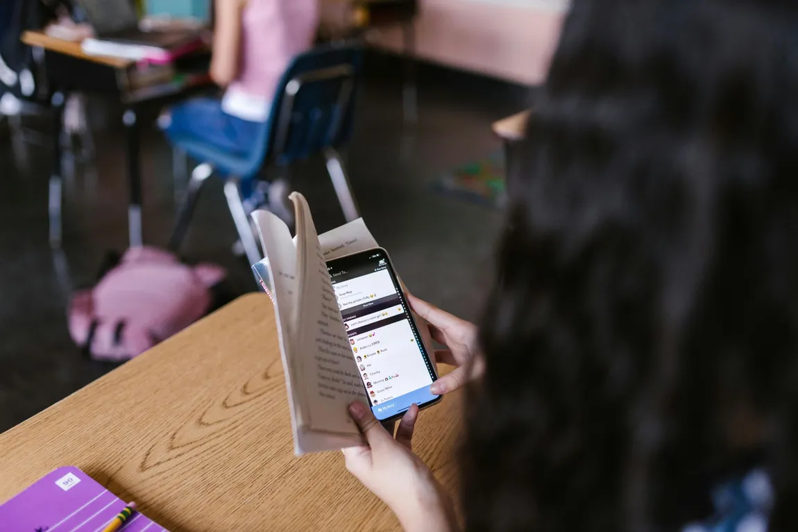 A student at a classroom desk quietly uses a phone kept low behind a propped-open book